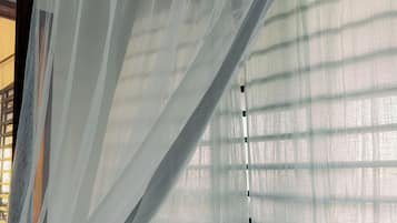 Una habitación con una cama cubierta por cortinas blancas translúcidas, una ventana con persianas horizontales y un cabecero de madera.