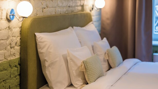 A neatly made bed with white linens and multiple pillows, a green upholstered headboard, and two wall-mounted light fixtures. - East-West Boutique Hotel Budapest (Budapest)