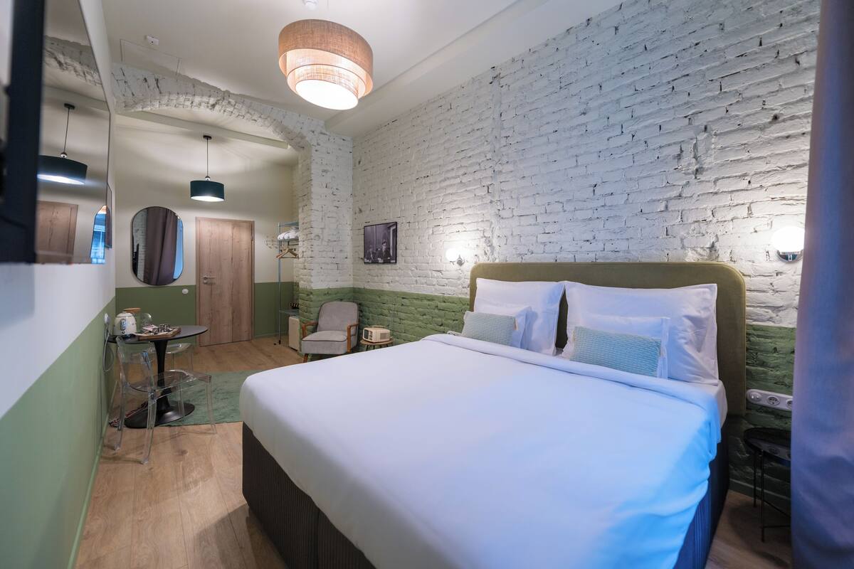 A hotel room with a large bed, a small table, a chair, a mirror, and a brick wall.
