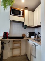 A compact kitchen with a microwave, a small sink, a wooden stool, and a dish rack.
