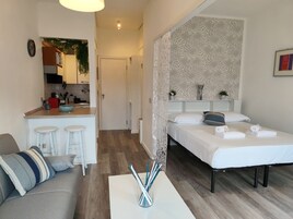 A modern hotel room with a kitchenette, a bed with white linens, a grey sofa, and a coffee table with pens.