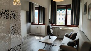 Uma sala de estar moderna com um sofá, uma mesa e um abajur. Há duas janelas com cortinas, um radiador e um painel decorativo na parede.