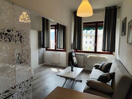 A modern living room with a sofa, a table, and a lamp. There are two windows with curtains, a radiator, and a decorative wall panel.