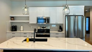 A modern kitchen with white cabinets, a stainless steel refrigerator, a built-in oven, a microwave, and pendant lighting.