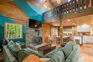 A cozy cabin interior with a stone fireplace, green upholstered sofas, a dining area, and a kitchen with white cabinets.