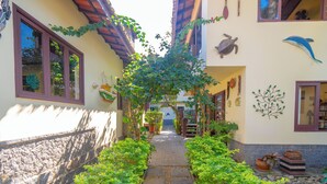 A narrow stone pathway flanked by lush greenery, leading to a building with a tiled roof and decorative wall art. - Vivenda do Flamboyant  (Ubatuba)