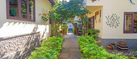 A narrow stone pathway flanked by lush greenery, leading to a building with a tiled roof and decorative wall art.