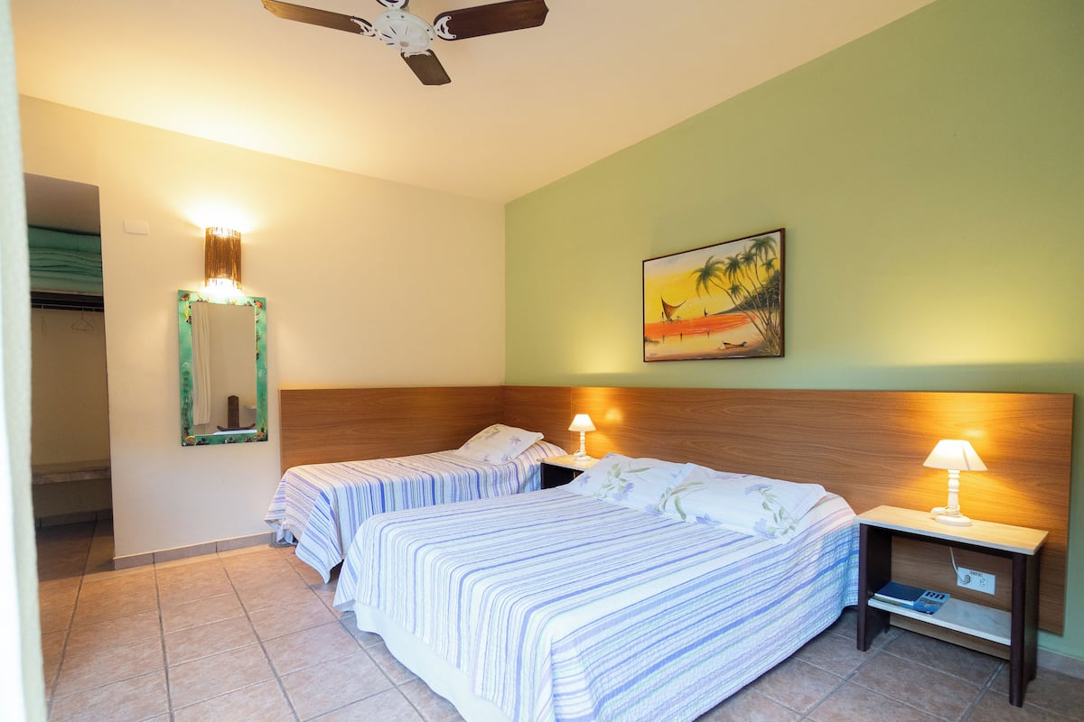 Habitación de hotel con dos camas, un ventilador de techo, una mesita de noche con lámpara, un cuadro en la pared y un baño visible a través de una puerta abierta.