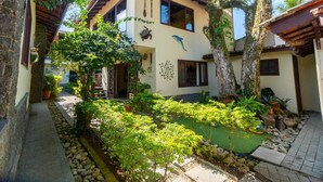 A two-story building with a tiled roof, a small garden with various plants, and a sign that reads "Duchas". - Vivenda do Flamboyant  (Ubatuba)