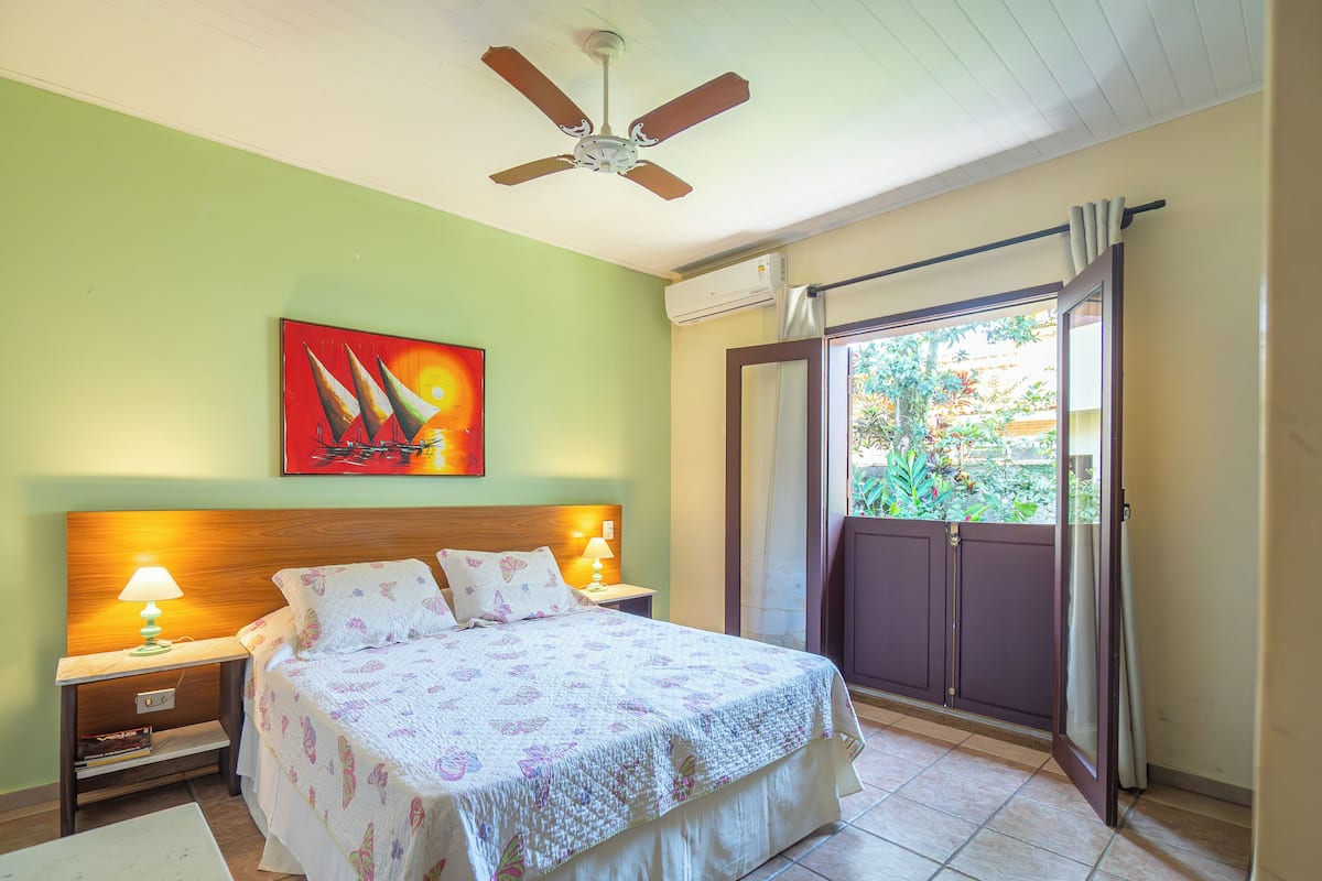 Un dormitorio con cama, dos lámparas de noche, ventilador de techo, ventana con cortinas y un cuadro en la pared.