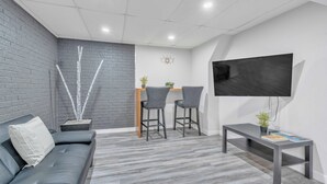 A modern living room with a grey sofa, two bar stools, a coffee table, a flat-screen TV, and a brick accent wall. - Luxury Basement Suite in Top Prime Location (Winnipeg)