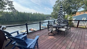Una terraza de madera con mobiliario exterior, una mesa y sillas, con vista a un lago tranquilo con barcos.