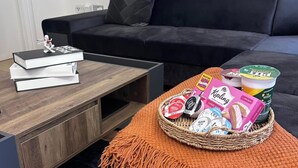 Un salón con un sofá oscuro, una mesa de centro de madera, y una cesta con aperitivos y libros.