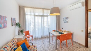 A modern living room with a sofa, dining table, and chairs. There is a large window with curtains, a hanging light fixture, and a wall-mounted air conditioner. - Bright beachfront flat (Bibione)