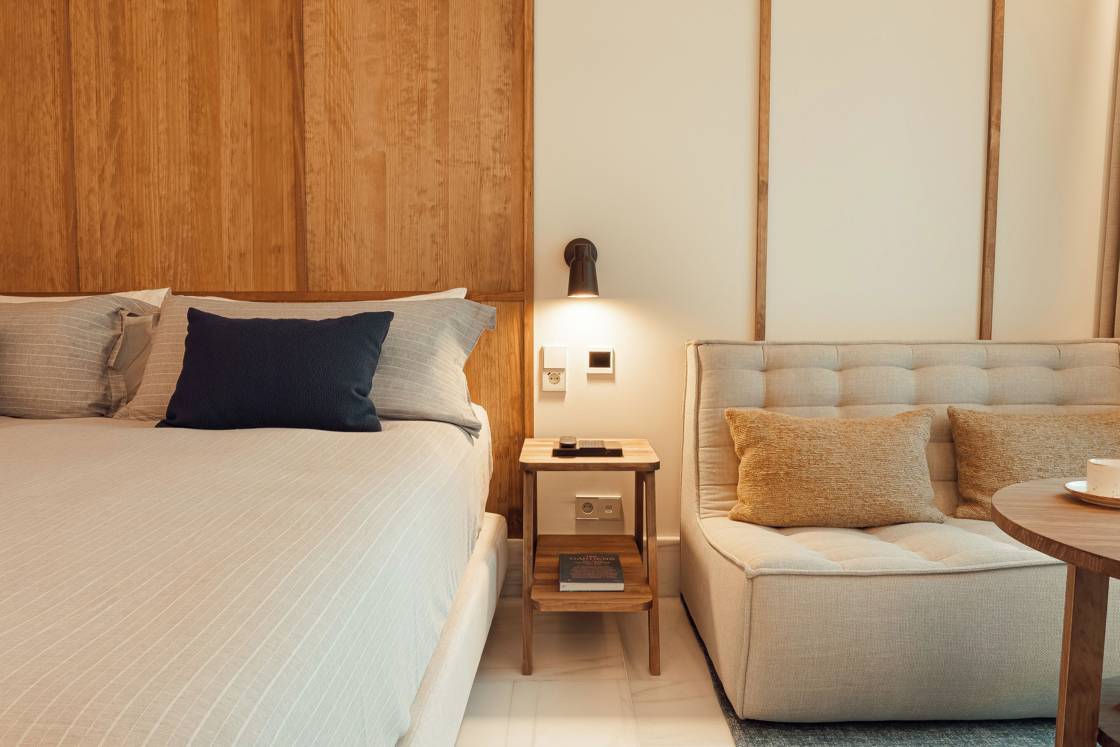 Hotellihuone, jossa on sänky, yöpöytä lampun kanssa, pieni jakkara ja sohva.