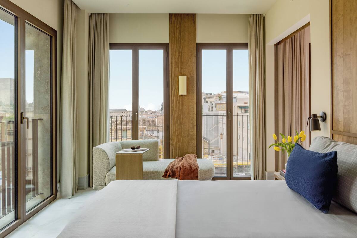 Habitación de hotel con una cama grande, un pequeño espacio para sentarse y vistas de la ciudad a través de amplios ventanales.