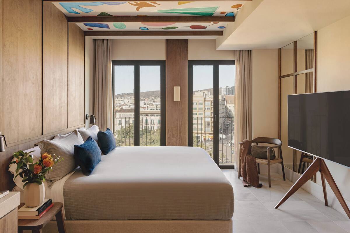 Ein Hotelzimmer mit einem großen Bett, einem Fernseher, einem Sessel und Blick auf die Stadt.