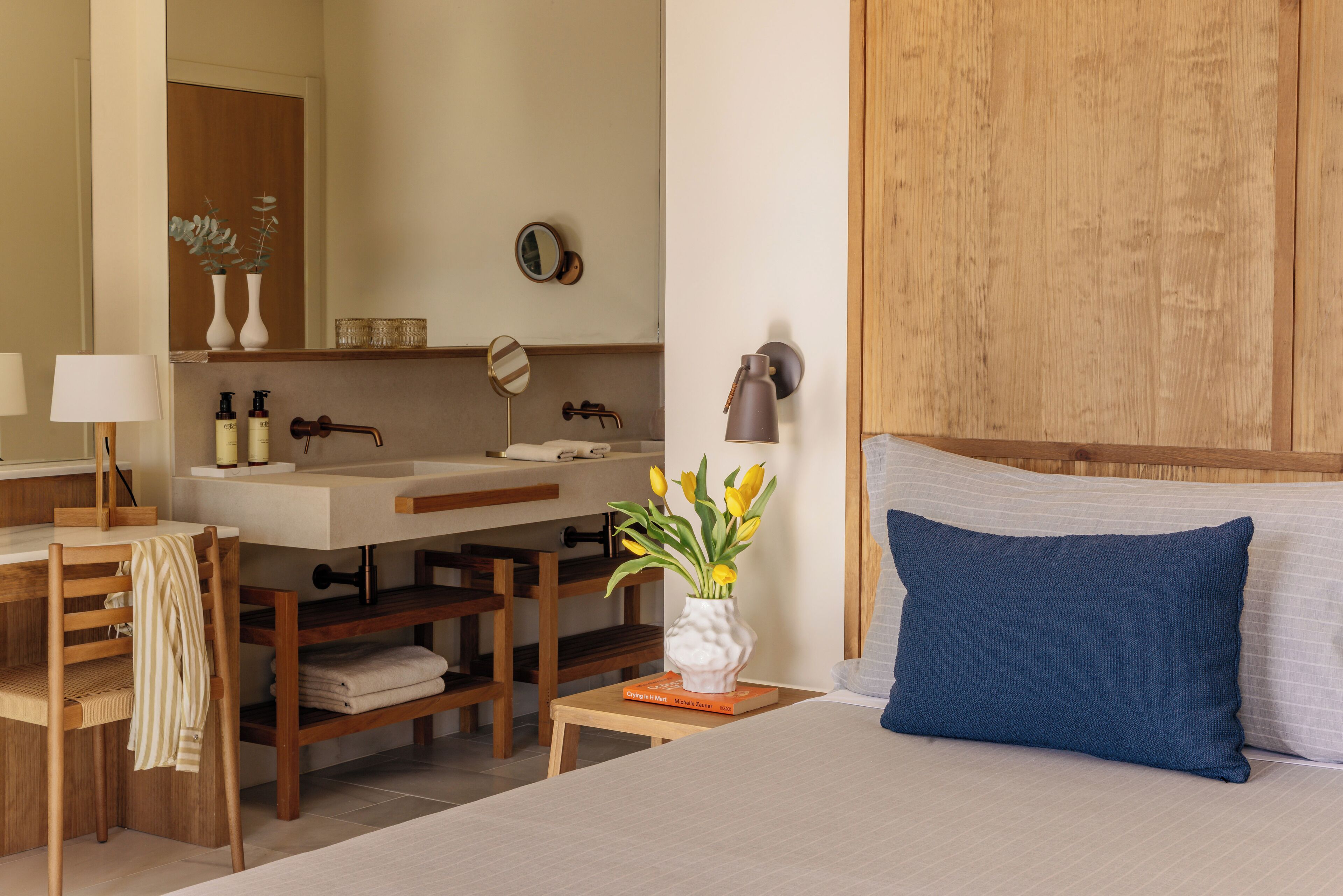 A bedroom with a bed, a nightstand, a bedside table with a vase of flowers, a wooden headboard, a wooden desk with a chair, a mirror, and a bathroom with a sink and towel rack.