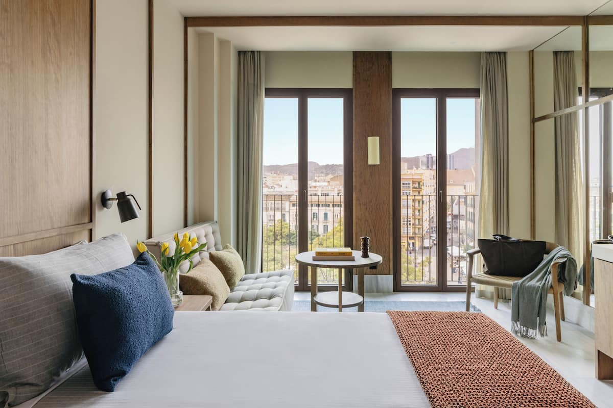 Habitación de hotel con una cama grande, una pequeña mesa redonda, una silla y vistas de la ciudad a través de grandes ventanales.