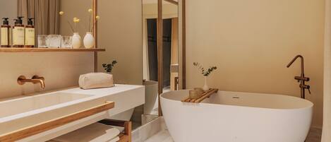 A modern bathroom with a freestanding bathtub, a large mirror, and a wooden vanity with multiple bottles and a vase.
