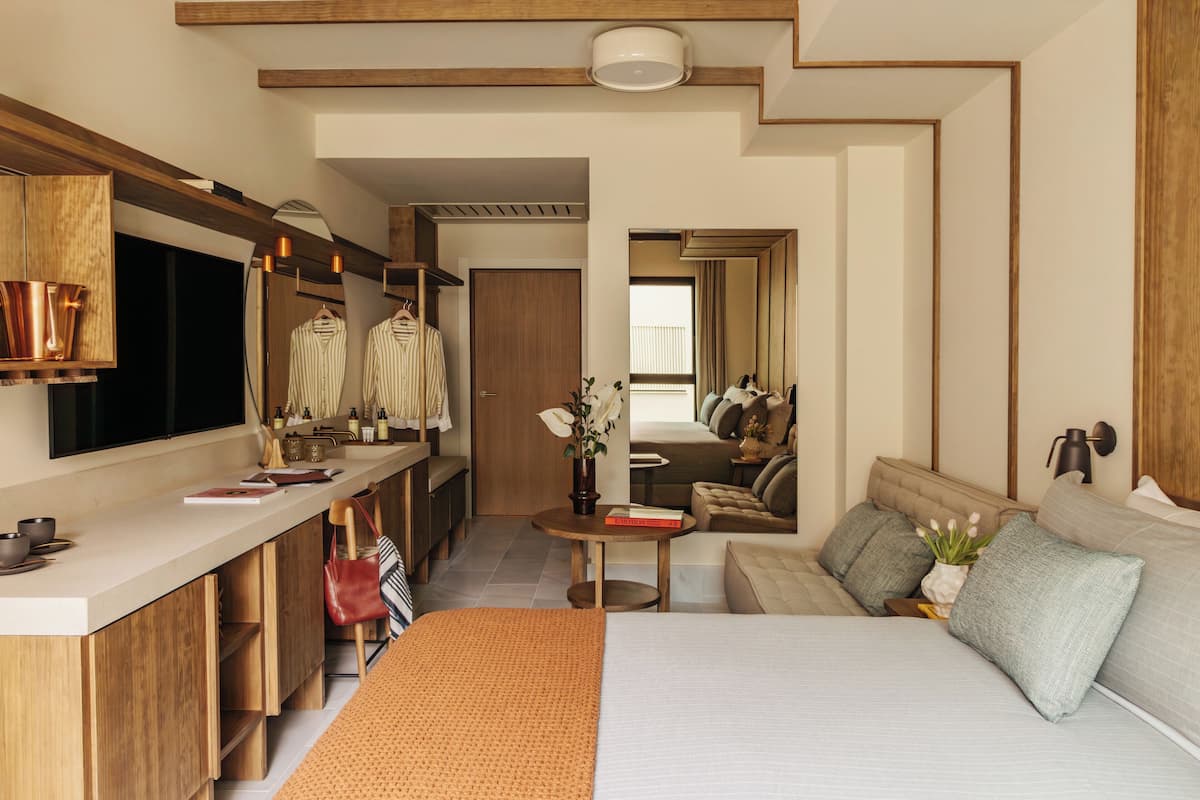 Una habitación de hotel moderna con una cama, un pequeño rincón de descanso, una kitchenette y un baño visible a través de una puerta abierta.