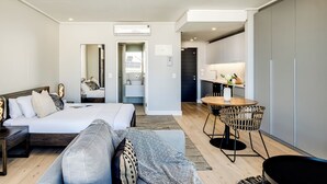 A modern hotel room with a bed, a sofa, a dining area with a table and chairs, a small kitchen, and a bathroom with a mirror and sink.