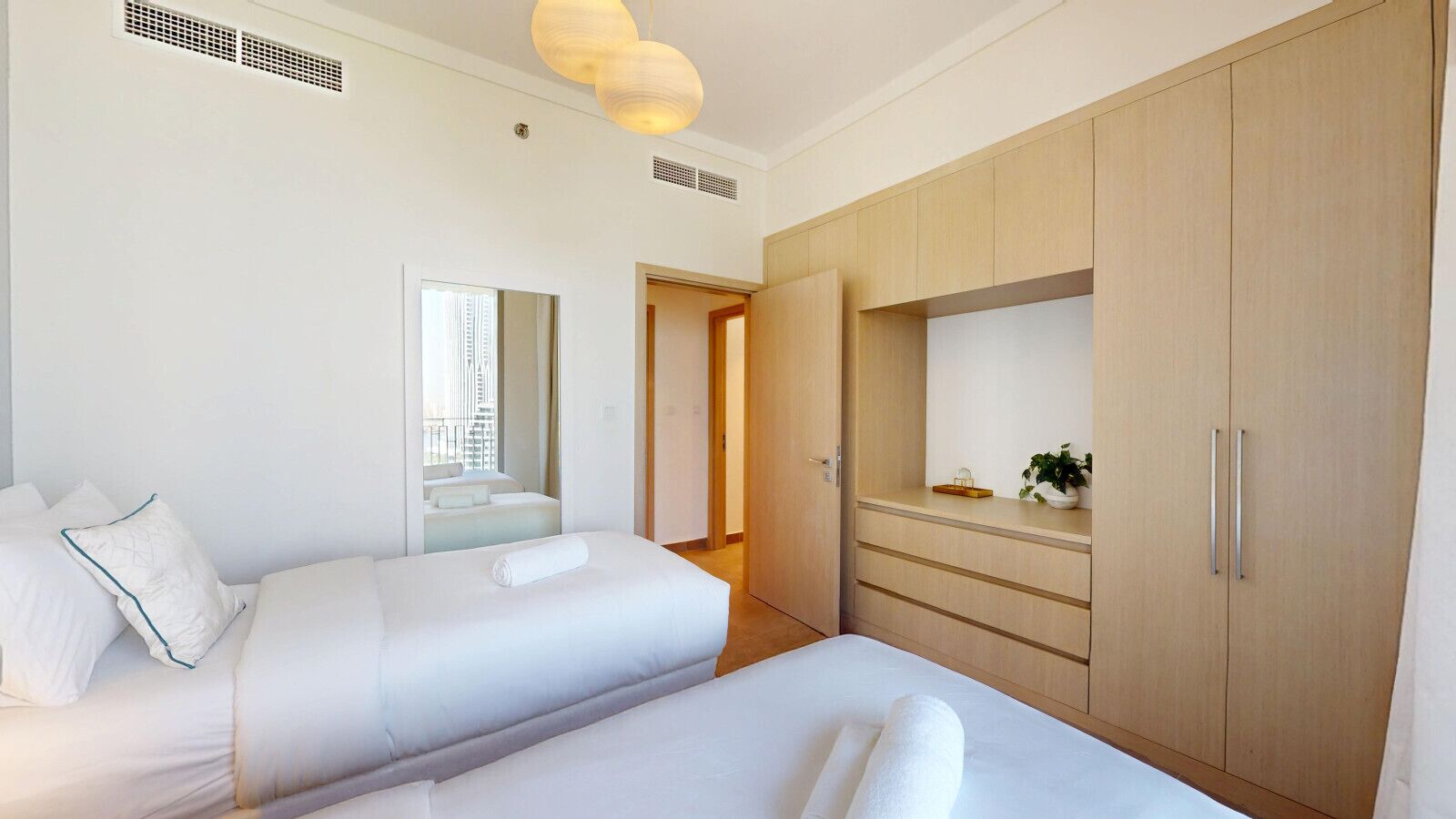 A hotel room with two beds, a wooden wardrobe, a mirror, and a small plant on the dresser.