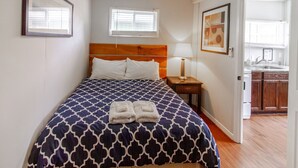 A hotel room with a bed, a nightstand with a lamp, a framed picture on the wall, and a view of the kitchen through an open door. - Couple`s Romantic PEC Getaway (Prince Edward)