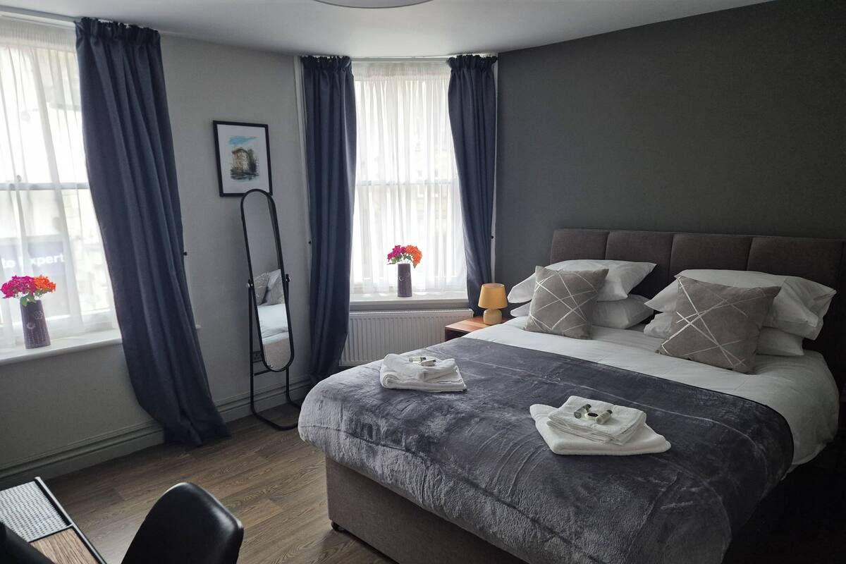 Una habitación de hotel moderna con una cama grande, una mesita de noche con lámpara, una silla, un espejo de pie y una ventana con cortinas.