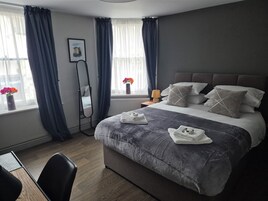 A modern hotel room with a large bed, a bedside table with a lamp, a chair, a standing mirror, and a window with curtains.