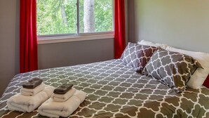 Un dormitorio con techo de madera, cama con ropa de cama estampada, cortinas rojas y ventana con vista a árboles.