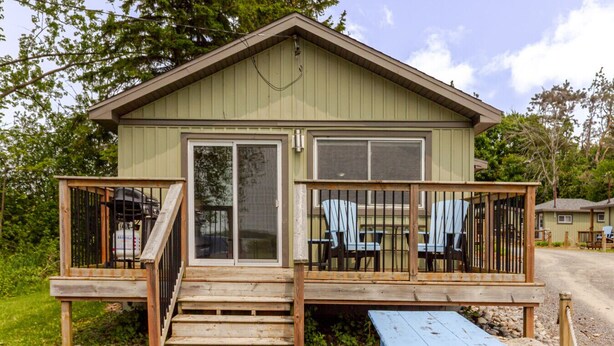 Una cabaña pequeña con una terraza de madera, dos puertas correderas de vidrio y un porche cubierto con sillas Adirondack.