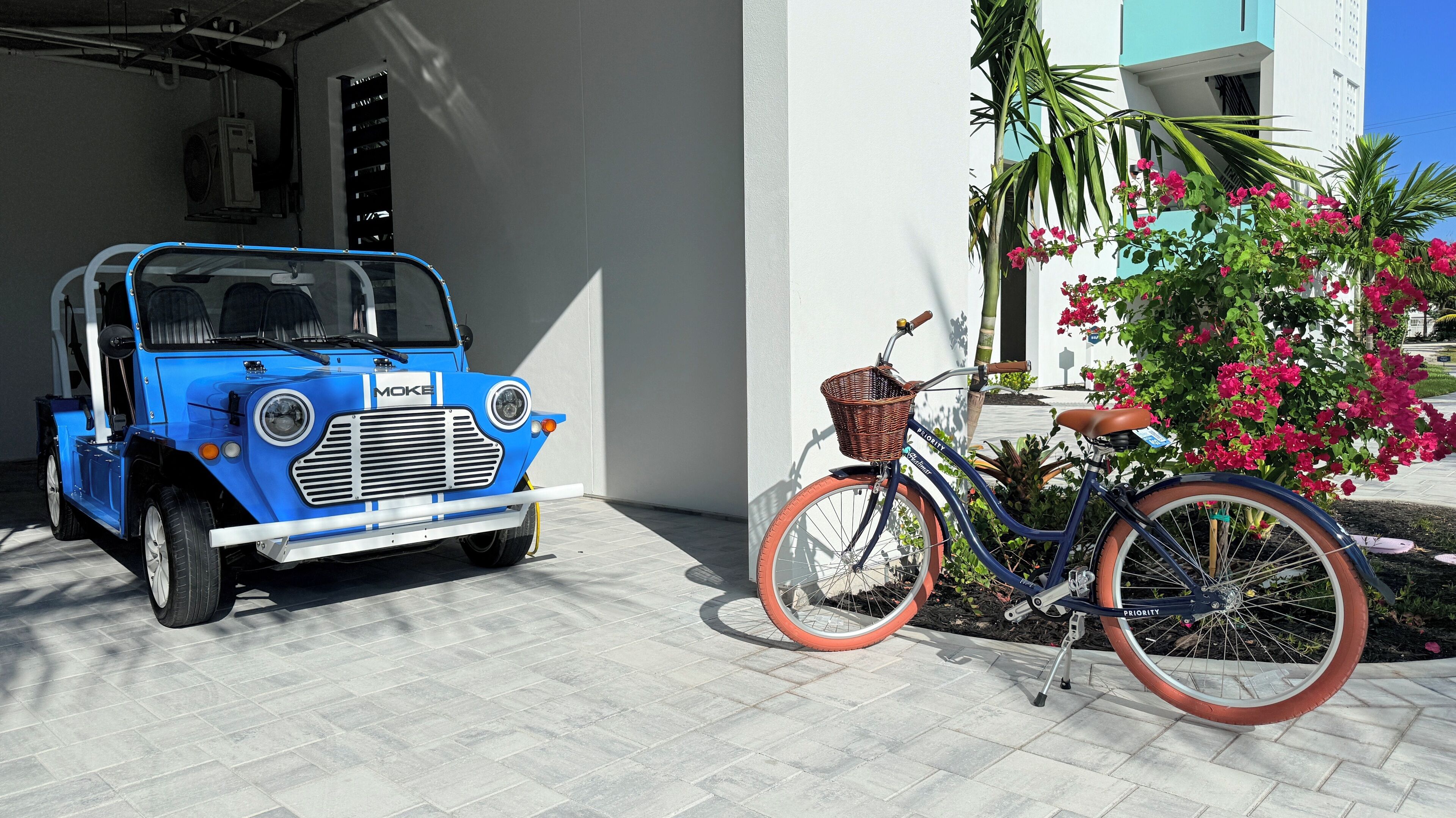 Une voiture bleue d’époque est garée dans un garage, à côté d’un vélo muni d’un panier.