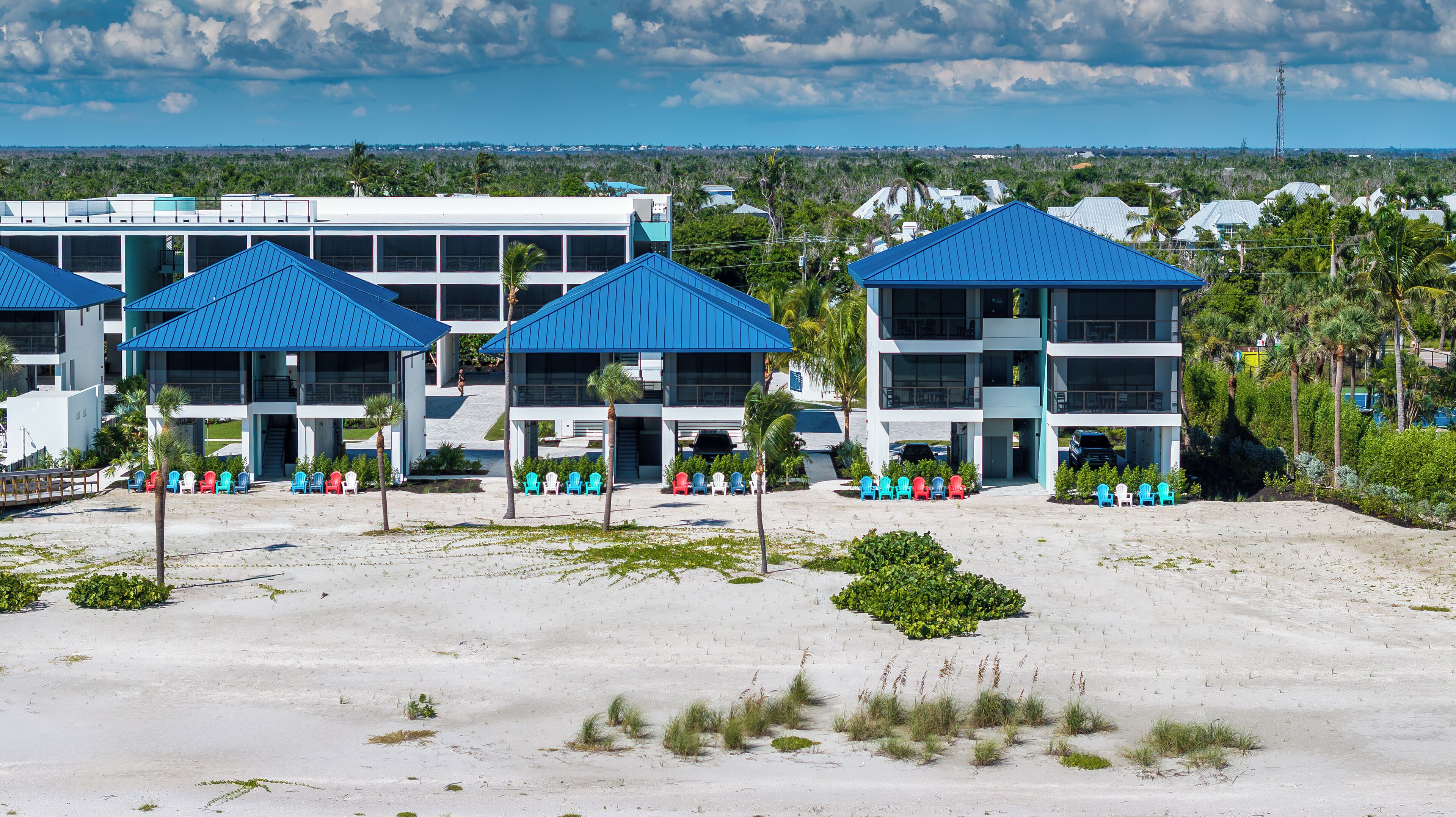 Un hotel fronte spiaggia con diversi edifici a tetti blu, una spiaggia sabbiosa e numerose sedie da spiaggia colorate.