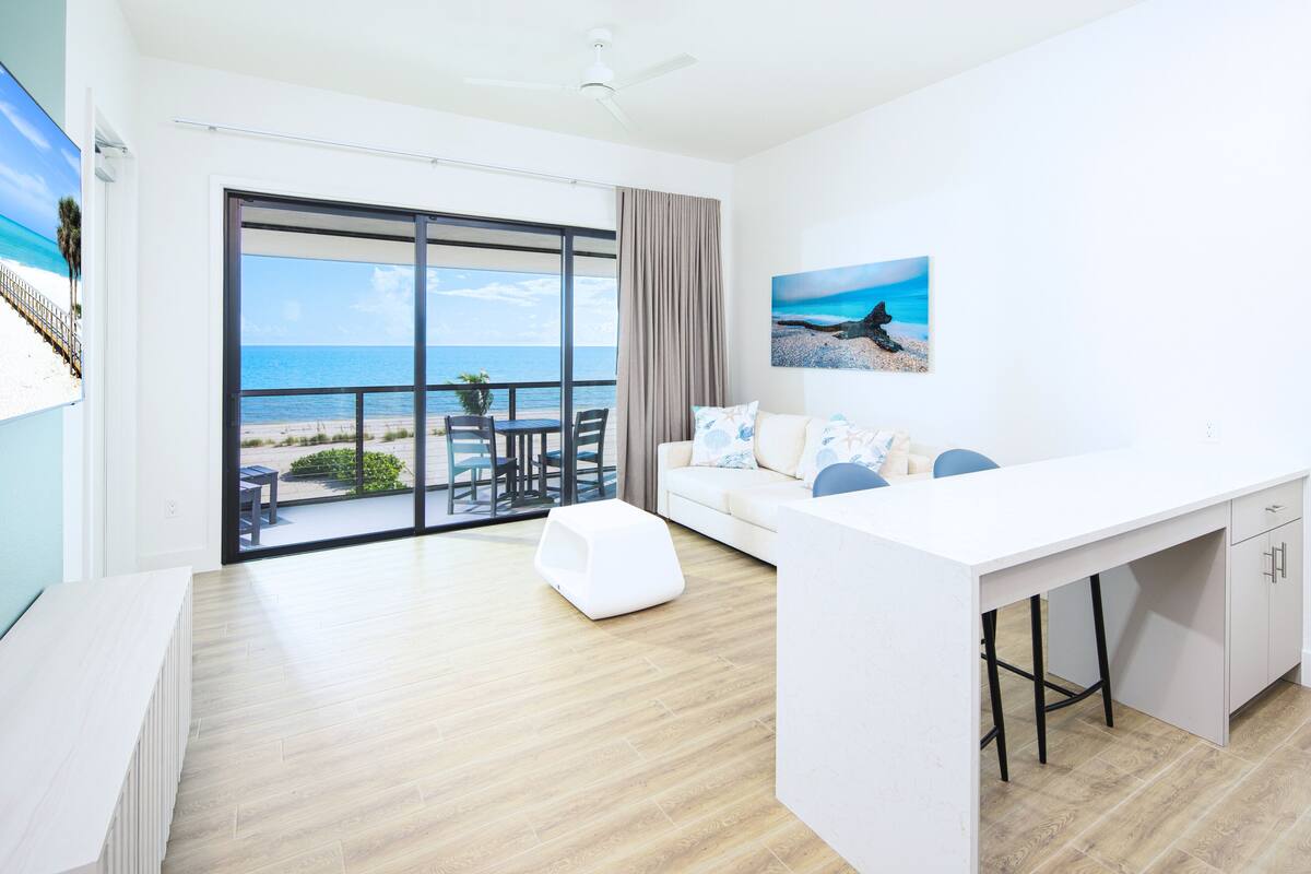 A modern room with a large sliding glass door leading to a balcony with a view of the beach. There is a white sofa, a small table, and a white desk with a chair.