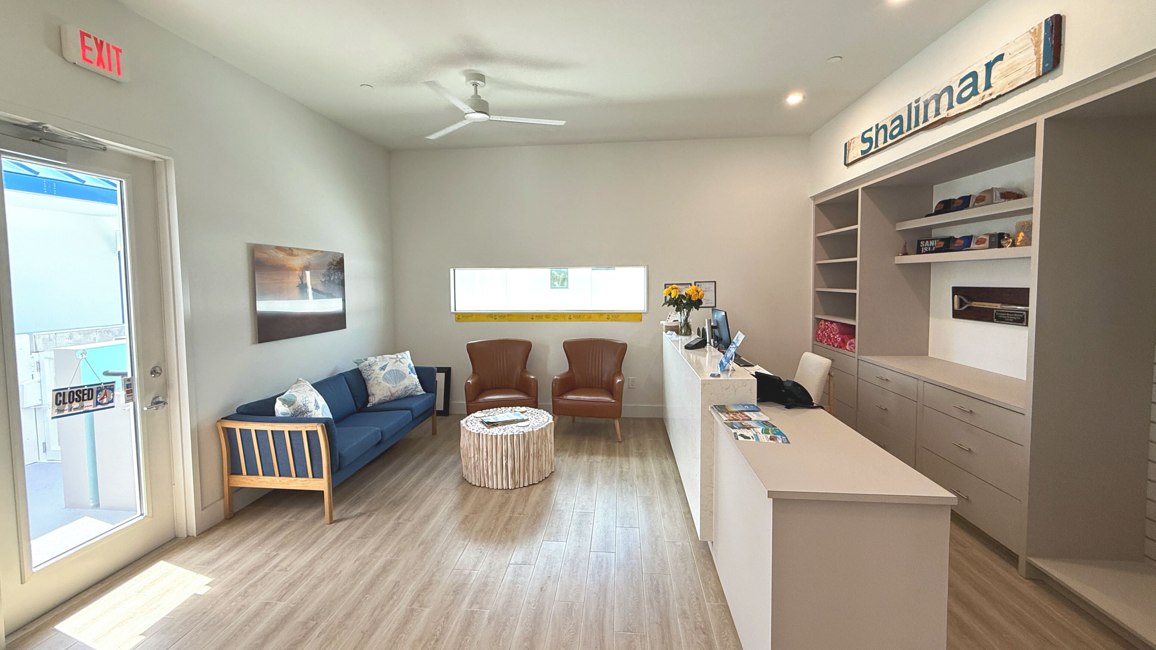 A reception area with a reception desk, two brown leather chairs, a blue sofa with patterned cushions, a small round table, and a ceiling fan.