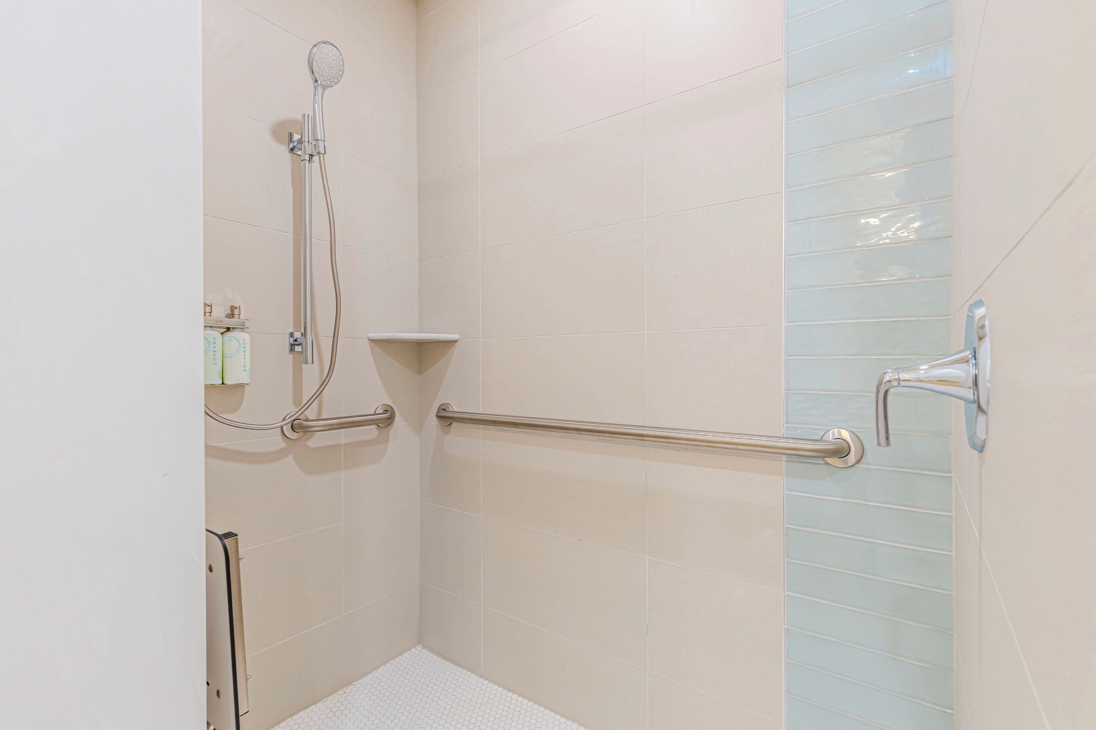 Un espace douche équipé d’un pommeau de douche à main, de barres d’appui et d’un sol carrelé de carreaux blancs.