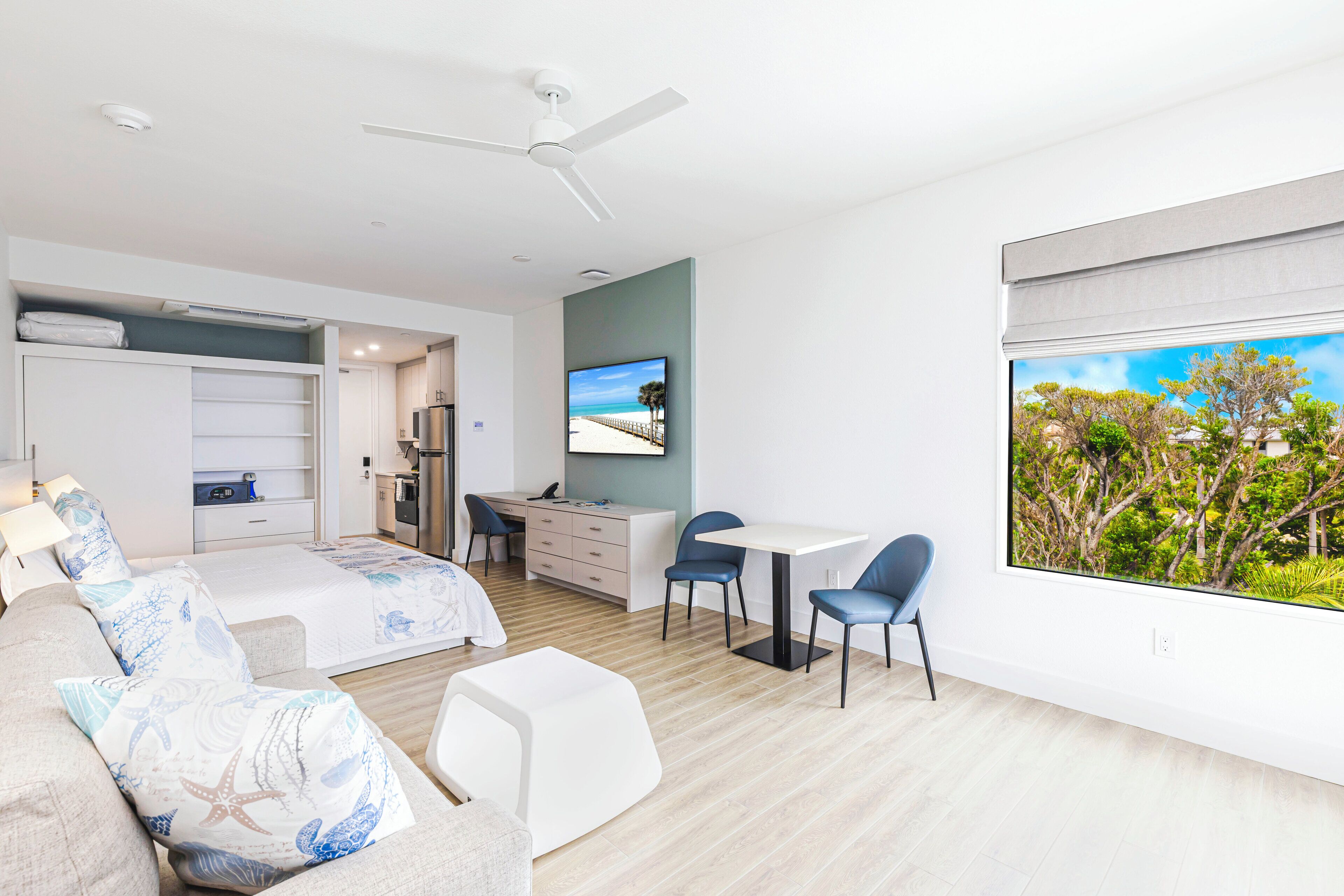 Une chambre d’hôtel moderne équipée d’un lit, d’un bureau, d’une chaise, d’une petite table et d’une grande fenêtre donnant sur un paysage arboré.