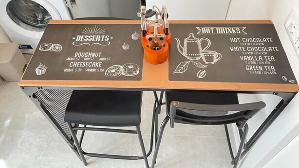 A kitchen area with a chalkboard menu listing desserts and hot drinks, a coffee machine, and a washing machine in the background.