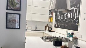 Une cuisine moderne avec des meubles blancs, une cuisinière à gaz, un évier et un tableau noir sur lequel est écrit « Bienvenue ». On peut voir des photos encadrées au mur, une plante et une assiette remplie de pâtisseries.