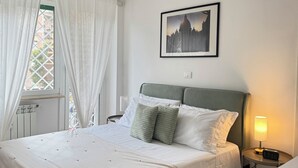 Une chambre à coucher bien rangée, avec un lit, une lampe de chevet, un tableau encadré au mur et une fenêtre avec des rideaux.