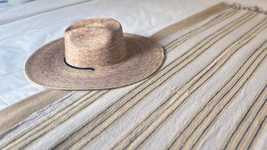 A bed with a straw hat on a striped pillow, a lamp, and framed artwork on the wall.