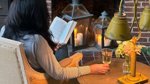 A woman reading a book at a wooden table with a fireplace in the background, surrounded by decorative items and candles.