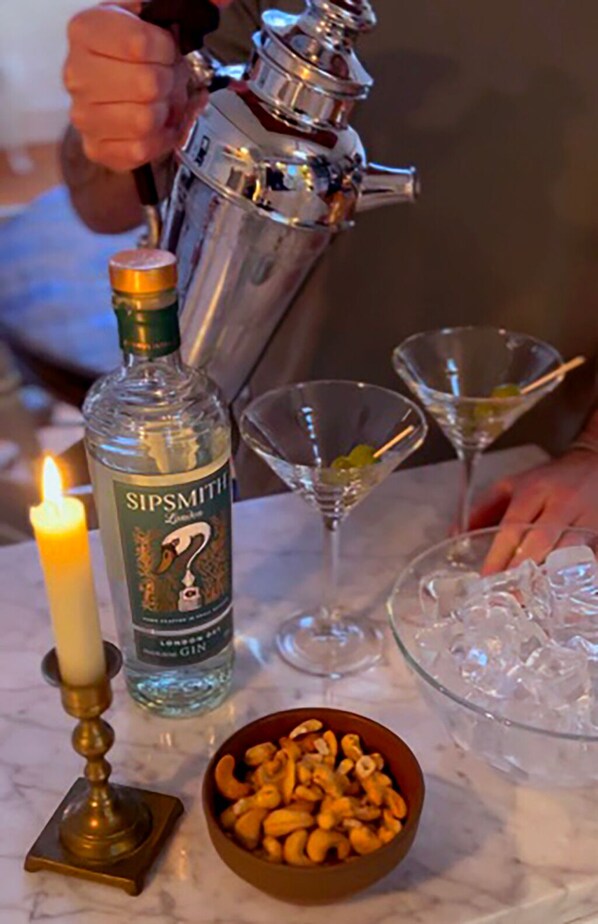 Un coctelera, una botella de ginebra Sipsmith, dos copas de martini, una vela y un bol de anacardos sobre una superficie de mármol.