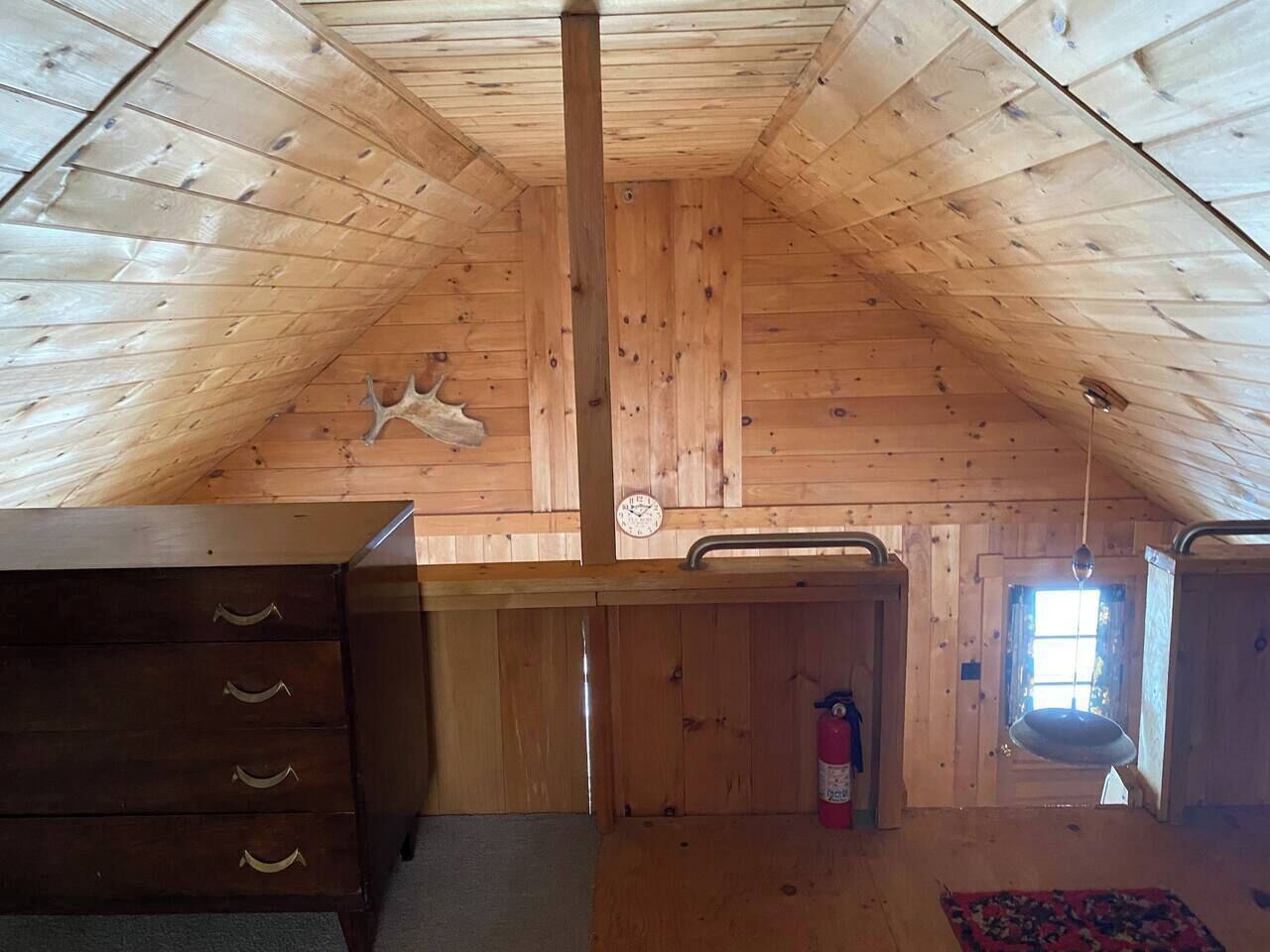 Une pièce mansardée en bois, avec un plafond en pente, une commode en bois, une fenêtre offrant une vue et un luminaire suspendu.