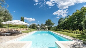 Ein rechteckiger Pool mit Steinrand, umgeben von einer Steinterrasse. Am Poolufer stehen Liegestühle und Sonnenschirme.