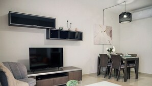 Ett modernt vardagsrum med en platt-TV, en soffa, ett soffbord med växter, en matplats med stolar och ett bord samt en hängande lampa.