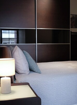 Una cama bien hecha con edredón blanco y varios almohadones, una lámpara de noche y un cabecero oscuro con diseño de cuadrícula.