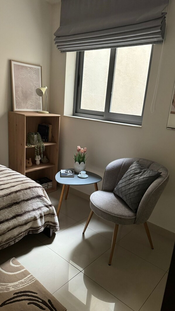 Un dormitorio moderno con una cama, una silla, una mesita con un jarrón de flores, una estantería y una ventana con persianas.