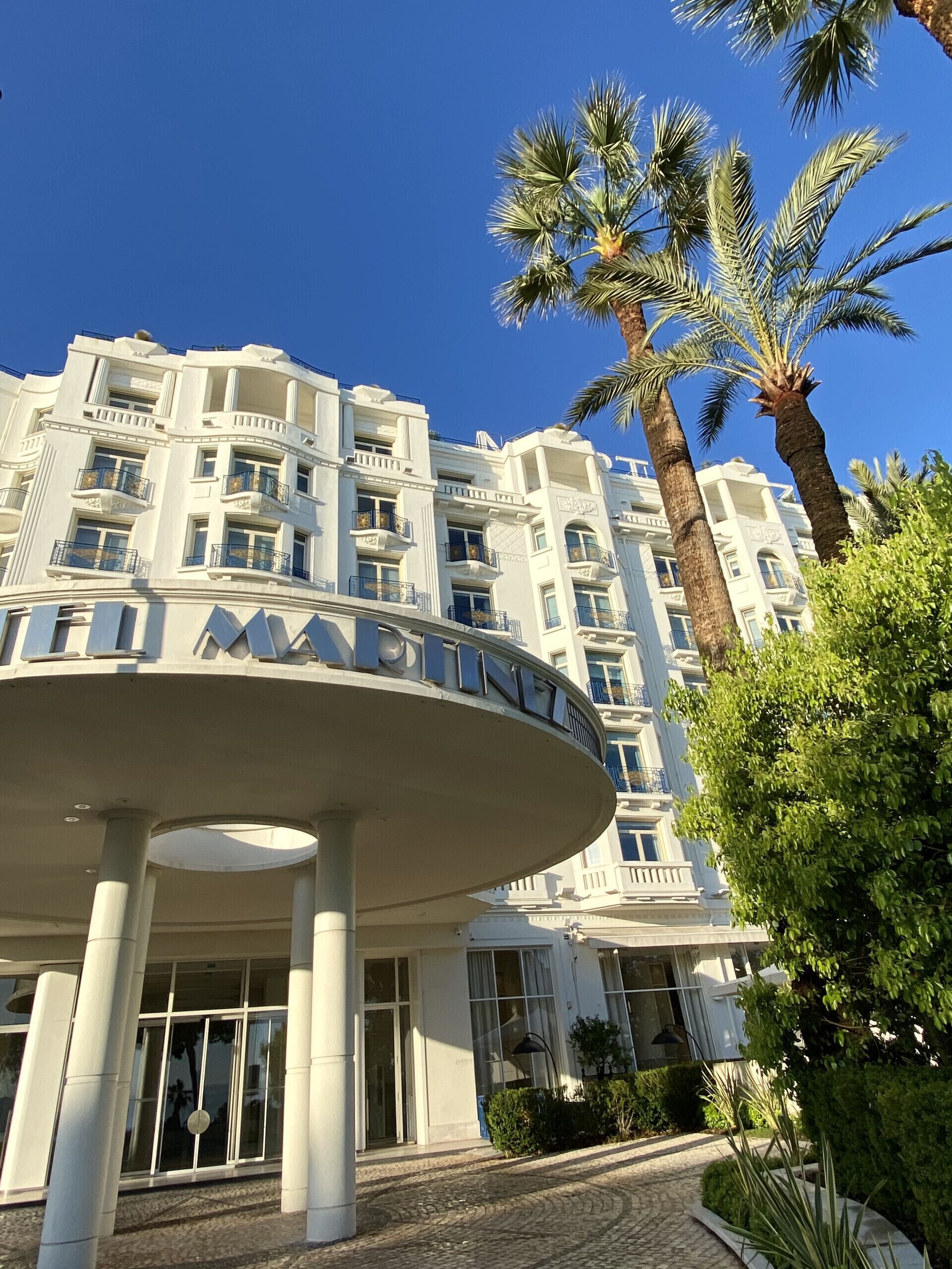 A hotel with a white facade, multiple balconies, and palm trees in the foreground.